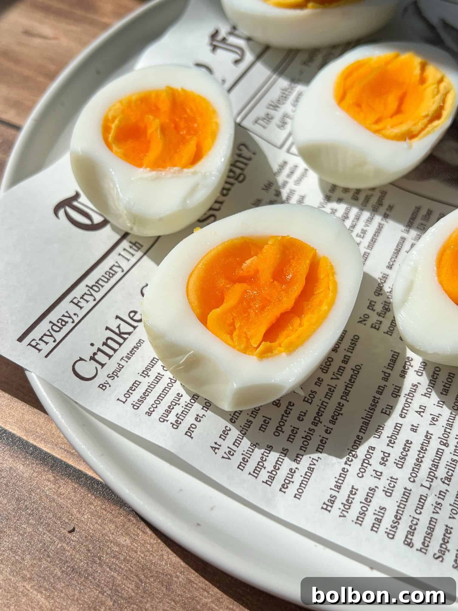 Close-up of halved soft-boiled eggs with bright orange yolks on a white plate lined with a newspaper-style paper, sitting on a wooden surface.