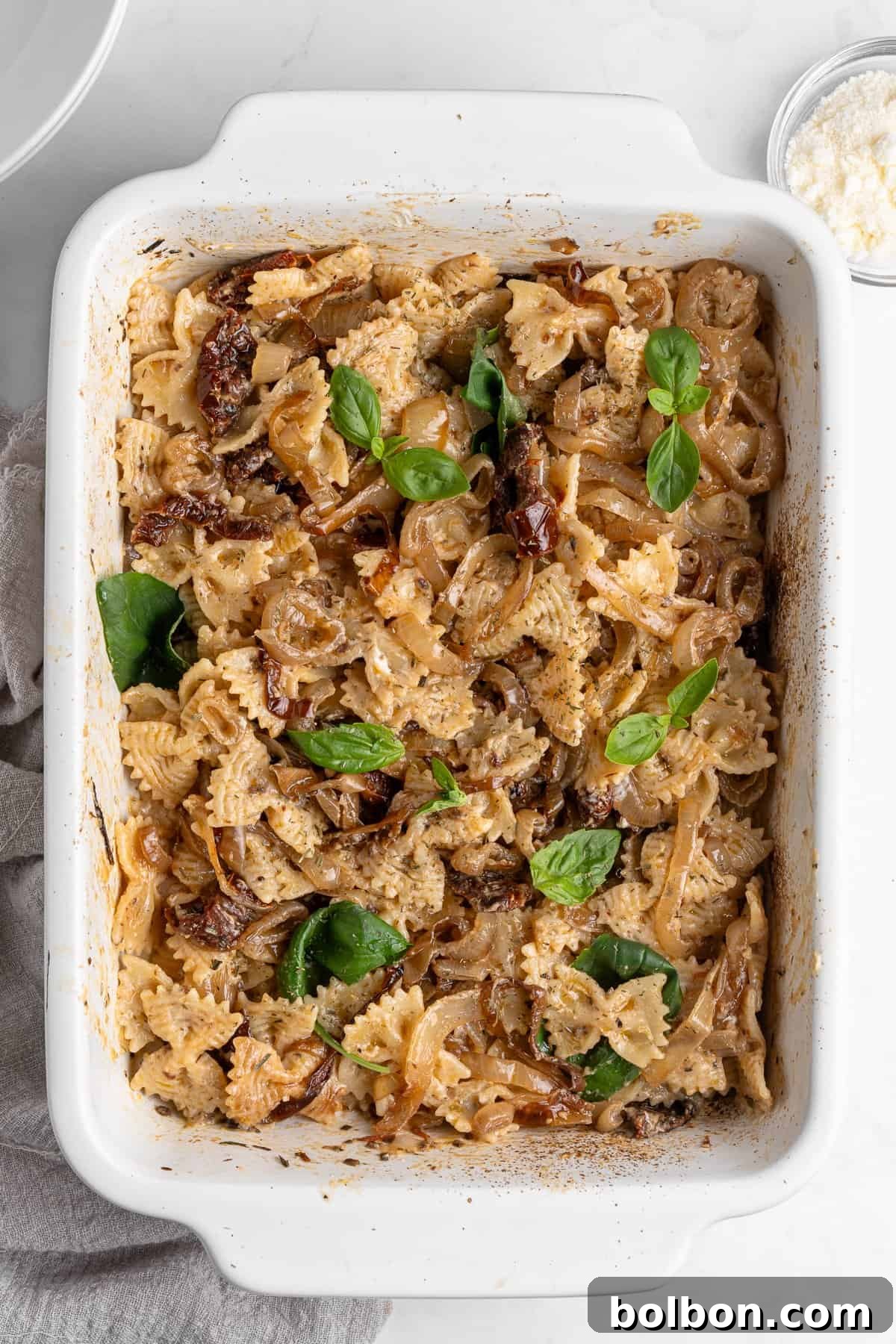 Slow-Cooked Onion Pasta 2 A delectable bowl of bow-tie pasta generously coated in a rich, golden caramelized onion sauce, adorned with vibrant sun-dried tomatoes and fresh basil leaves, finished with a delicate sprinkle of grated Parmesan cheese. A small, elegant bowl of Parmesan sits nearby, inviting an extra garnish.