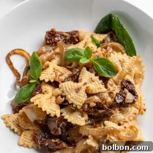 Slow-Cooked Onion Pasta 11 A white bowl filled with pasta topped with caramelized onions, sun-dried tomatoes, and fresh basil leaves. A small dish with grated cheese is in the background, along with basil sprigs on a light surface.