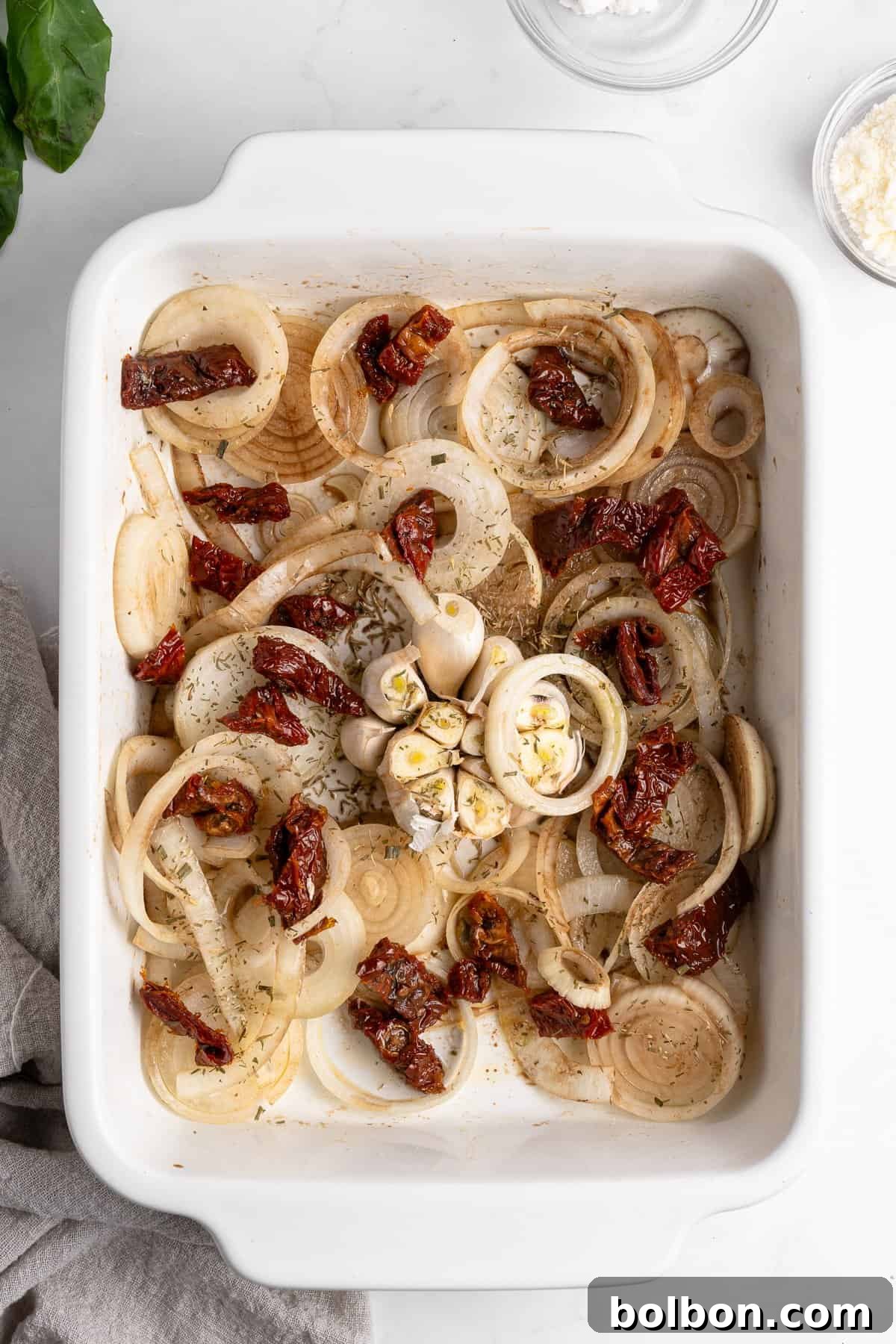 Slow-Cooked Onion Pasta 4 A pristine white baking dish, prepared and ready for the oven, showcasing neatly layered onion slices, bright sun-dried tomatoes, and a strategically halved bulb of garlic, all generously seasoned with a blend of aromatic herbs. The dish rests on a white marble surface, partially surrounded by a soft beige cloth and small, elegant bowls containing additional ingredients, hinting at the culinary journey ahead.