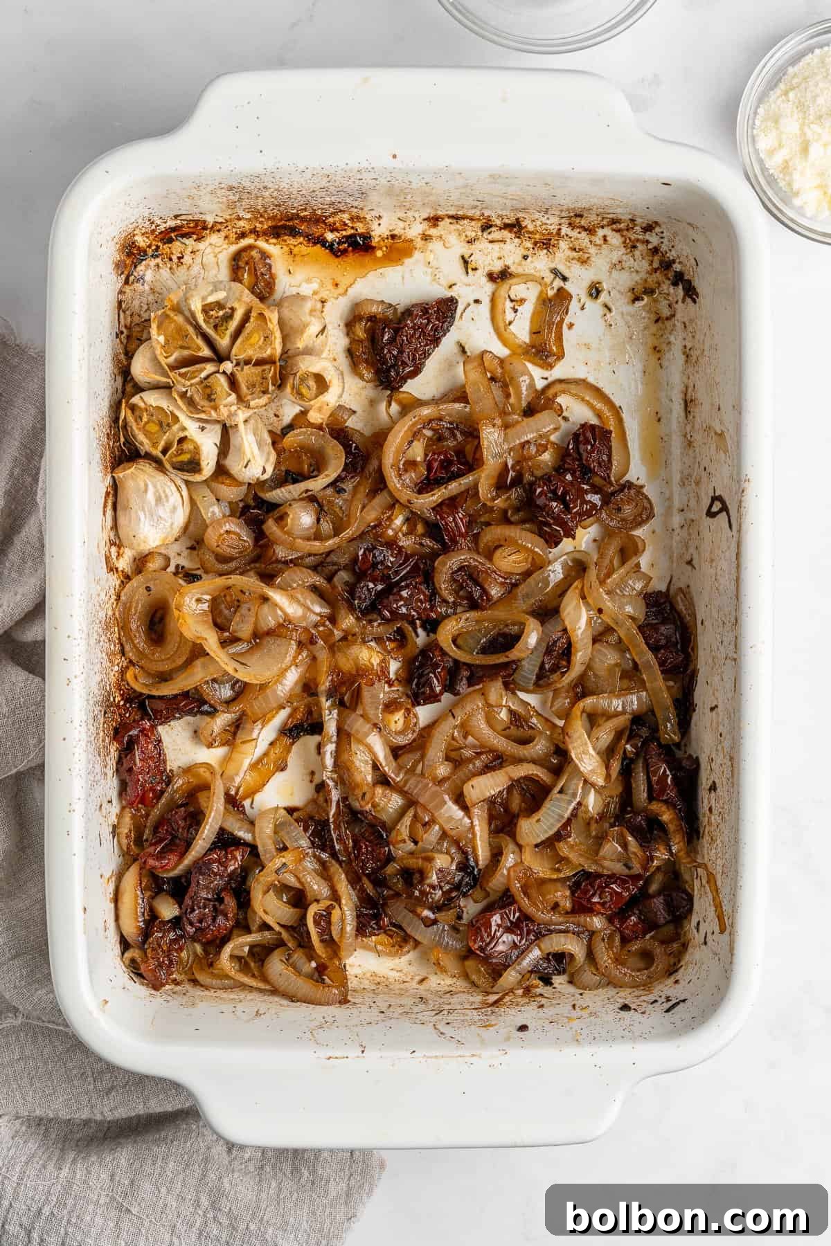 Slow-Cooked Onion Pasta 5 A white baking dish freshly removed from the oven, brimming with beautifully roasted onion slices, tender garlic cloves, and intensely flavored sun-dried tomatoes. The ingredients have achieved a rich, browned hue, with inviting areas of char on their edges, indicating perfect caramelization. A soft gray cloth peeks from the left side, ready to protect the surface.
