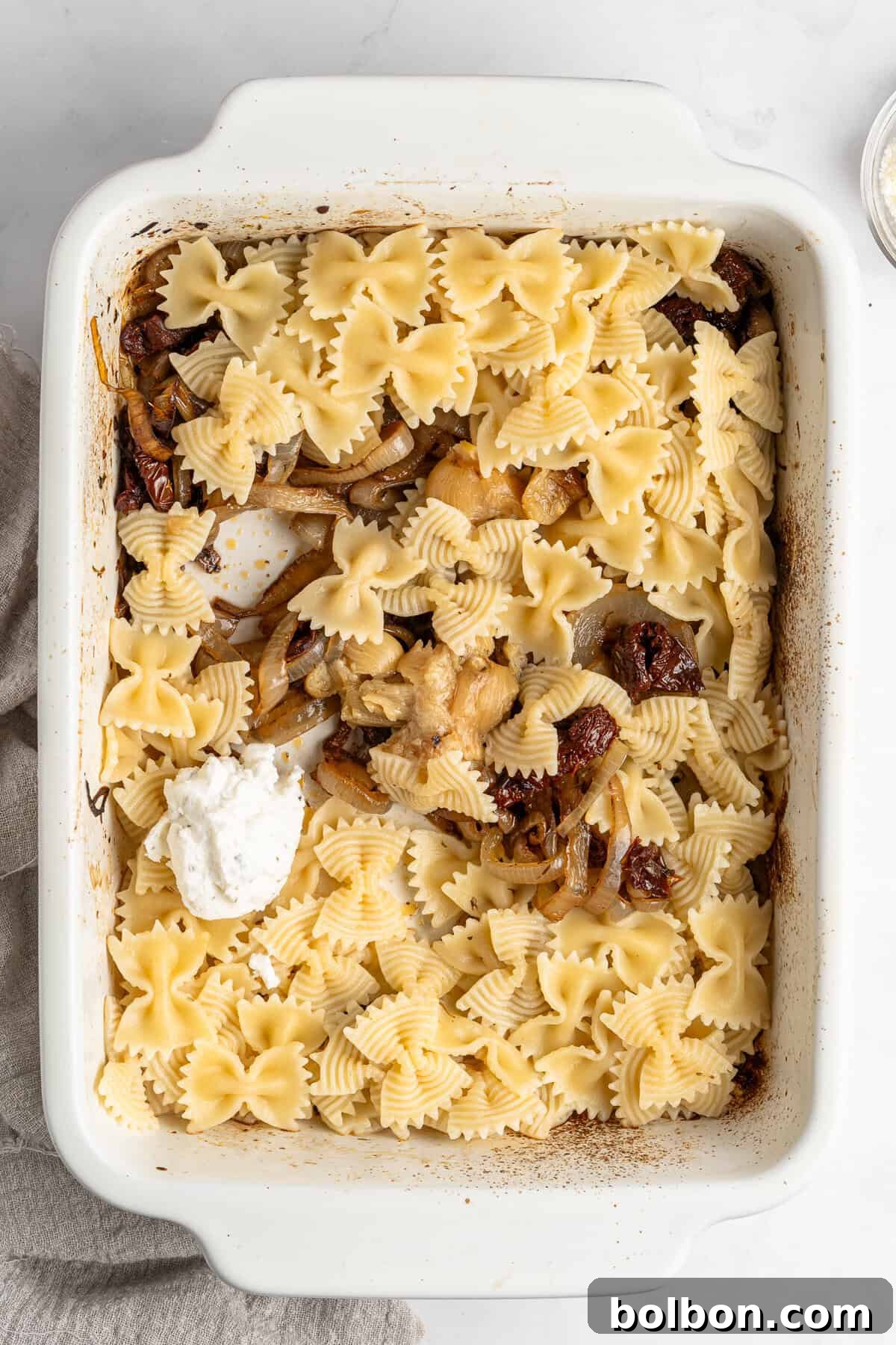 Slow-Cooked Onion Pasta 6 A tantalizing white baking dish filled with perfectly cooked bowtie pasta, generously mixed with tender sautéed onions and vibrant sun-dried tomatoes. A creamy dollop of ricotta cheese crowns the center, ready to melt into the dish. The edges of the dish show subtle browning, hinting at its oven-baked goodness. A soft beige cloth gracefully drapes along one side, enhancing the rustic charm.