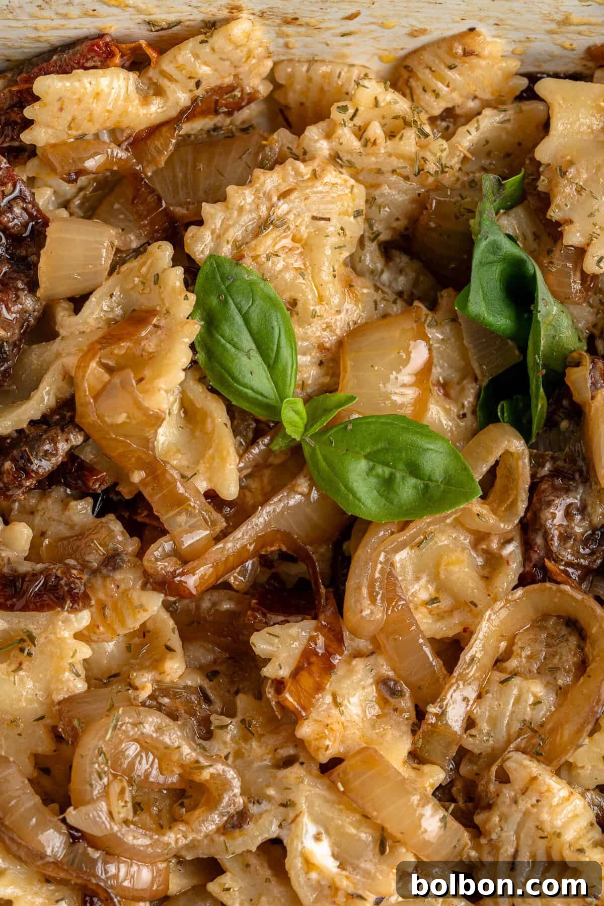 Slow-Cooked Onion Pasta 8 A close-up, appetizing view of a bow-tie pasta mixture, artfully garnished with vibrant fresh basil leaves. The dish exudes a creamy texture and is perfectly seasoned with a blend of aromatic herbs, promising a delightful culinary experience.