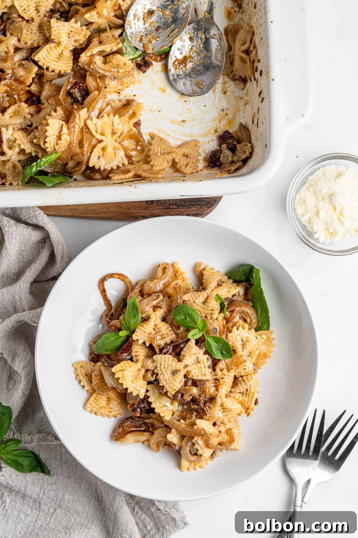 Slow-Cooked Onion Pasta 9 A beautifully composed white plate showcasing bow-tie pasta meticulously mixed with deeply caramelized onions, earthy mushrooms, and vibrant sun-dried tomatoes, exquisitely garnished with fresh basil. Beside it rests a matching baking dish filled with more of the delightful pasta, accompanied by two elegant spoons, a soft napkin, a small bowl of finely grated cheese, and two ready forks, all set for a perfect meal.