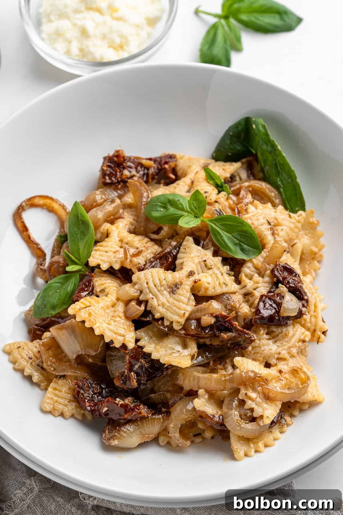Slow-Cooked Onion Pasta 10 A charming white bowl filled with a generous serving of pasta, luxuriously topped with golden caramelized onions, vibrant sun-dried tomatoes, and fresh, aromatic basil leaves. In the background, a small elegant dish holds grated cheese, accompanied by sprigs of basil artfully arranged on a light surface, creating an inviting and picturesque scene.