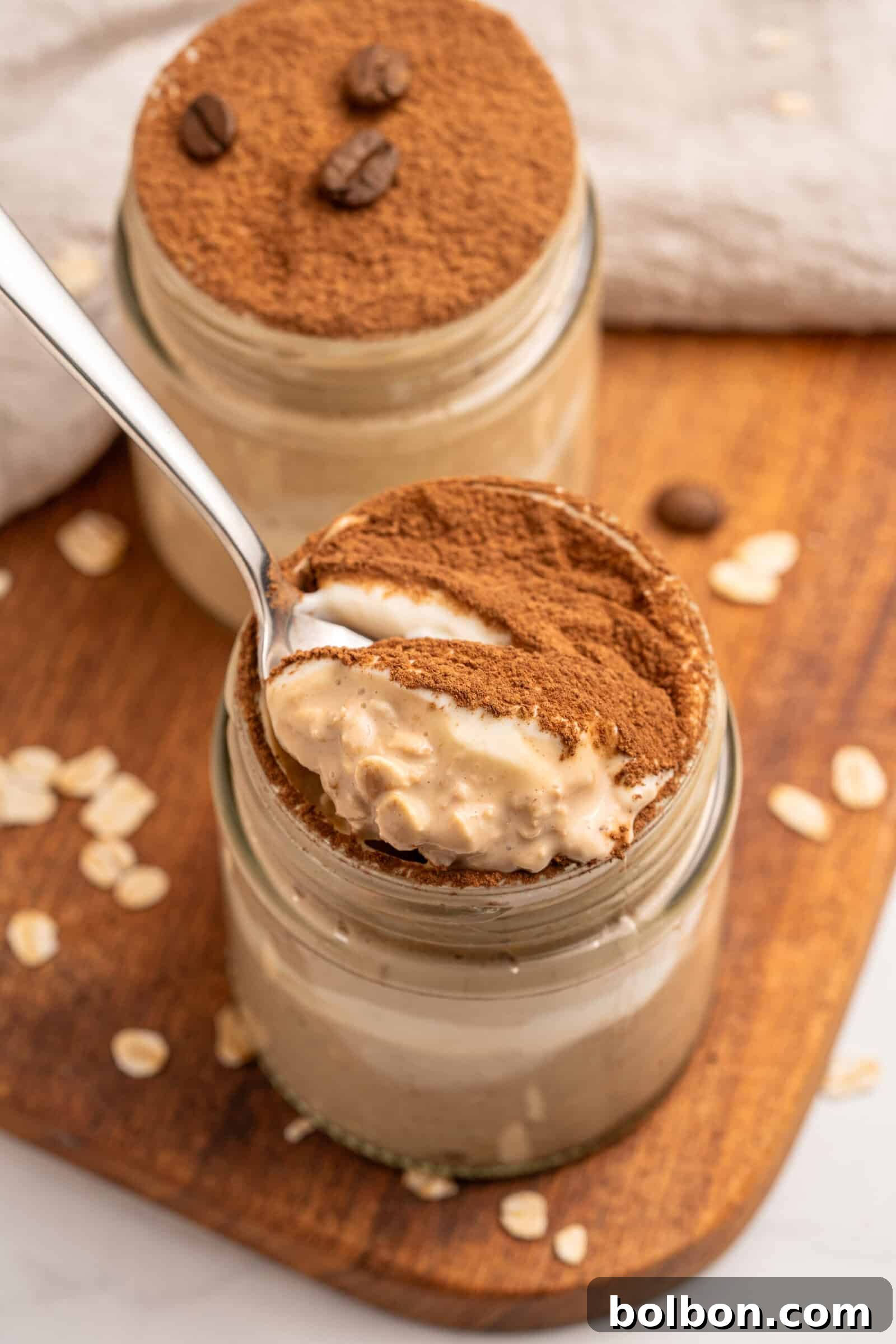 Dreamy Tiramisu Overnight Oats 2 A jar of creamy dessert topped with cocoa powder, with a spoon scooping out a portion. Another jar sits behind it, both on a wooden board sprinkled with oats and coffee beans.