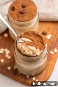 Dreamy Tiramisu Overnight Oats 5 A jar of creamy dessert topped with cocoa powder, with a spoon scooping out a portion. Another jar sits behind it, both on a wooden board sprinkled with oats and coffee beans.