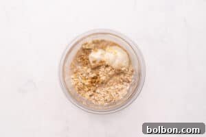 Dreamy Tiramisu Overnight Oats 3 A glass bowl containing dry oats, water, and a dollop of yogurt, viewed from above on a white surface.