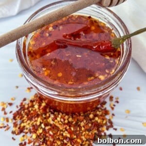 A glass jar filled with chili-infused honey, topped with a whole dried chili and a honey dipper resting on the rim. Crushed red pepper flakes are scattered around the jar on a white surface.