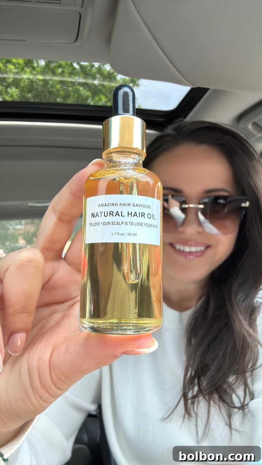 A woman with dark hair and sunglasses holds up a bottle of "Natural Hair Oil" with a dropper lid inside a car, smiling as she displays the product toward the camera.