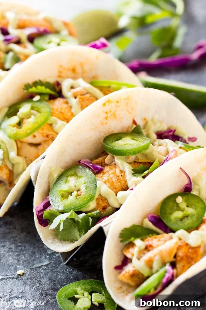Two vibrant Chipotle Lime Fish Tacos served on a rustic wooden board, garnished with fresh cilantro and a generous drizzle of creamy avocado crema. A bowl of crunchy jalapeno lime slaw is visible in the background.