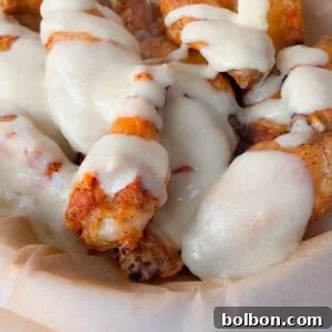 Alfredo Chicken Wings in a parchment-lined tray are being topped with a generous drizzle of creamy white sauce.