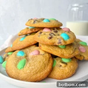 A plate of chocolate chip cookies topped with pastel-colored candy-coated chocolates, made using our Apple Cookies Recipe, sits beside a glass of milk in the background.
