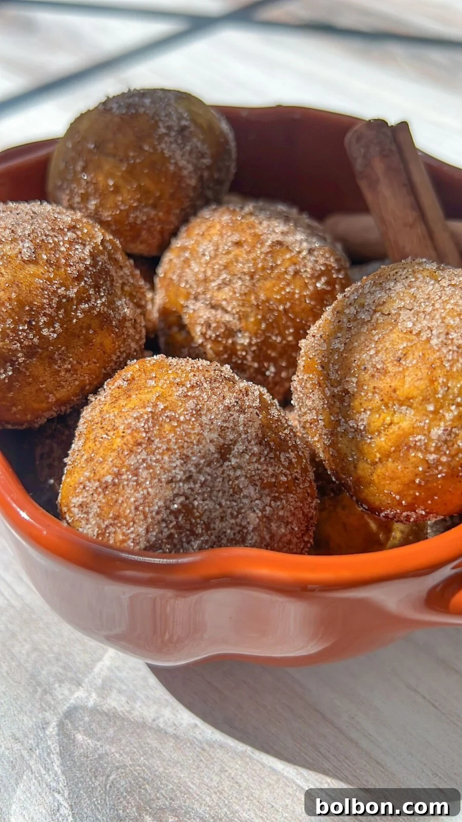 Quick 4 Ingredient Pumpkin Donut Bites 2 A close-up of pumpkin spice donut holes in an orange bowl. The donut holes are coated with cinnamon sugar, and two cinnamon sticks are nestled among them. The bowl is placed on a white wooden surface, and the sunlight casts soft shadows over the scene.