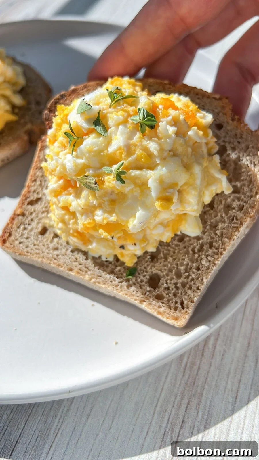 A hand holding a slice of toasted bread topped generously with creamy, golden baked feta eggs, garnished with fresh green herbs. The bread rests on a crisp white plate, set against a light wooden table, bathed in warm, inviting sunlight.