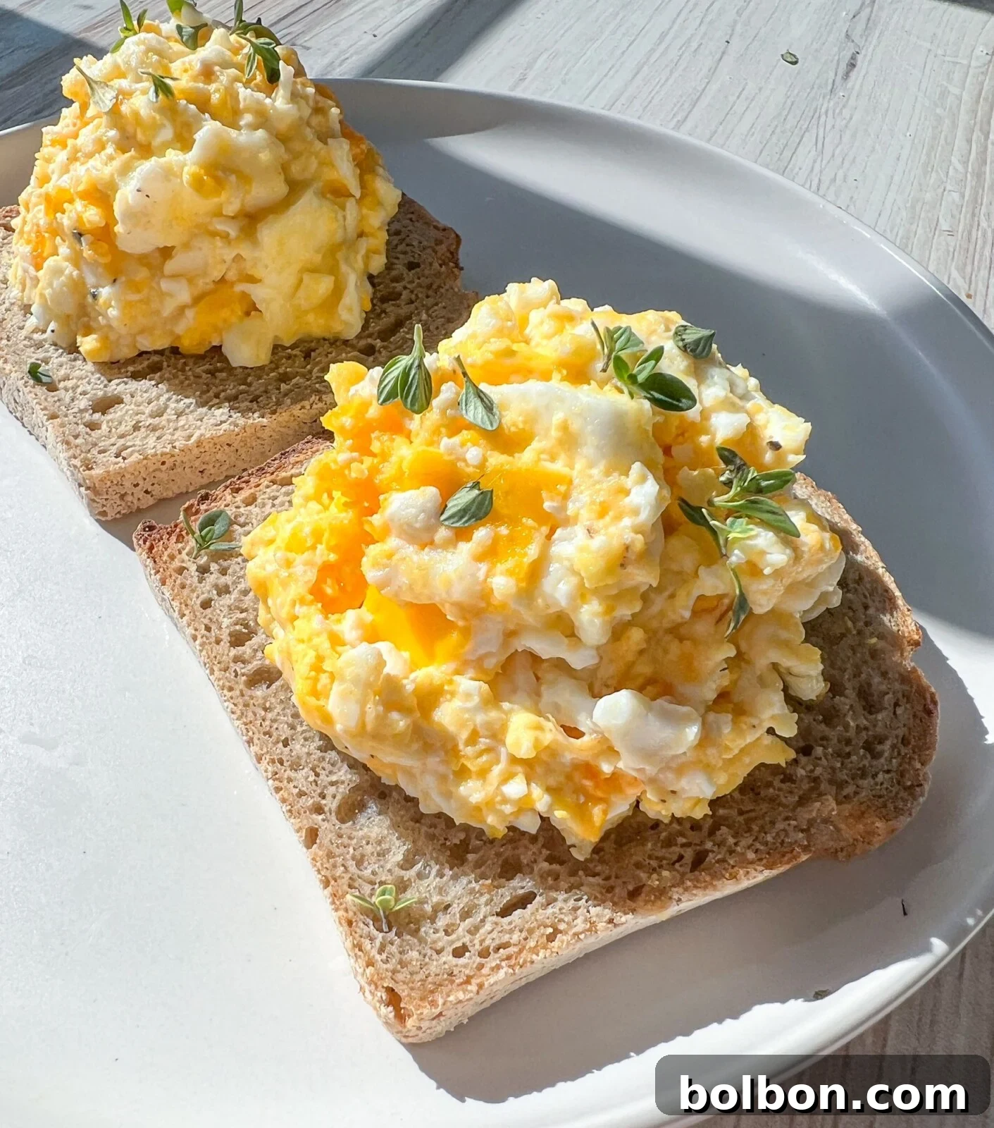 Two slices of golden-brown whole grain bread, generously topped with creamy baked feta eggs and delicately garnished with fresh thyme sprigs. The plate rests on a rustic light wooden surface, illuminated by natural sunlight, highlighting the appetizing textures.