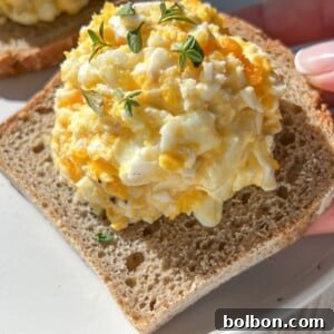 A hand holds a slice of bread topped with creamy egg salad and fresh herbs, over a white plate on a light table—perfect for pairing with 4 Ingredient Pumpkin Donut Holes for a delightful meal or snack.