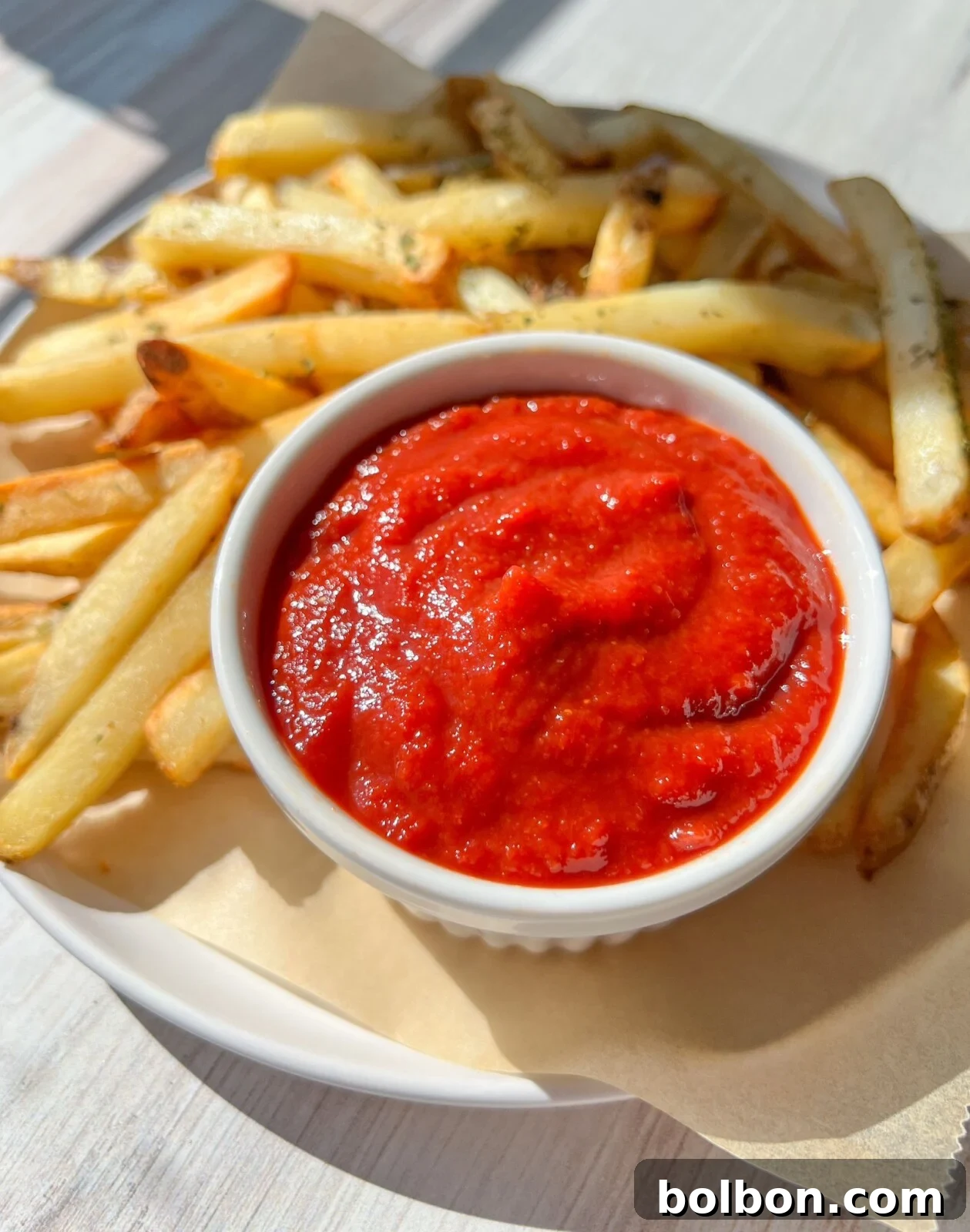 A close-up of a white plate filled with golden-brown french fries, garnished with fresh herbs. In the center of the plate is a small white bowl brimming with bright red homemade ketchup, ready for dipping. The plate rests on a pale wooden surface, bathed in natural sunlight, creating inviting shadows. This perfect pairing would also be wonderful with 4 Ingredient Pumpkin Donut Holes.