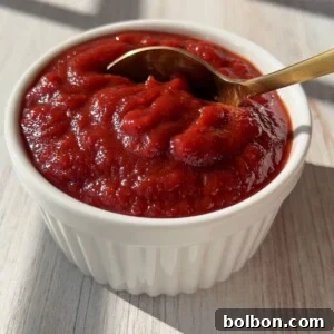 A white ramekin filled with thick, rich red homemade ketchup, with a gold spoon resting inside, sits on a light wooden surface in natural sunlight.