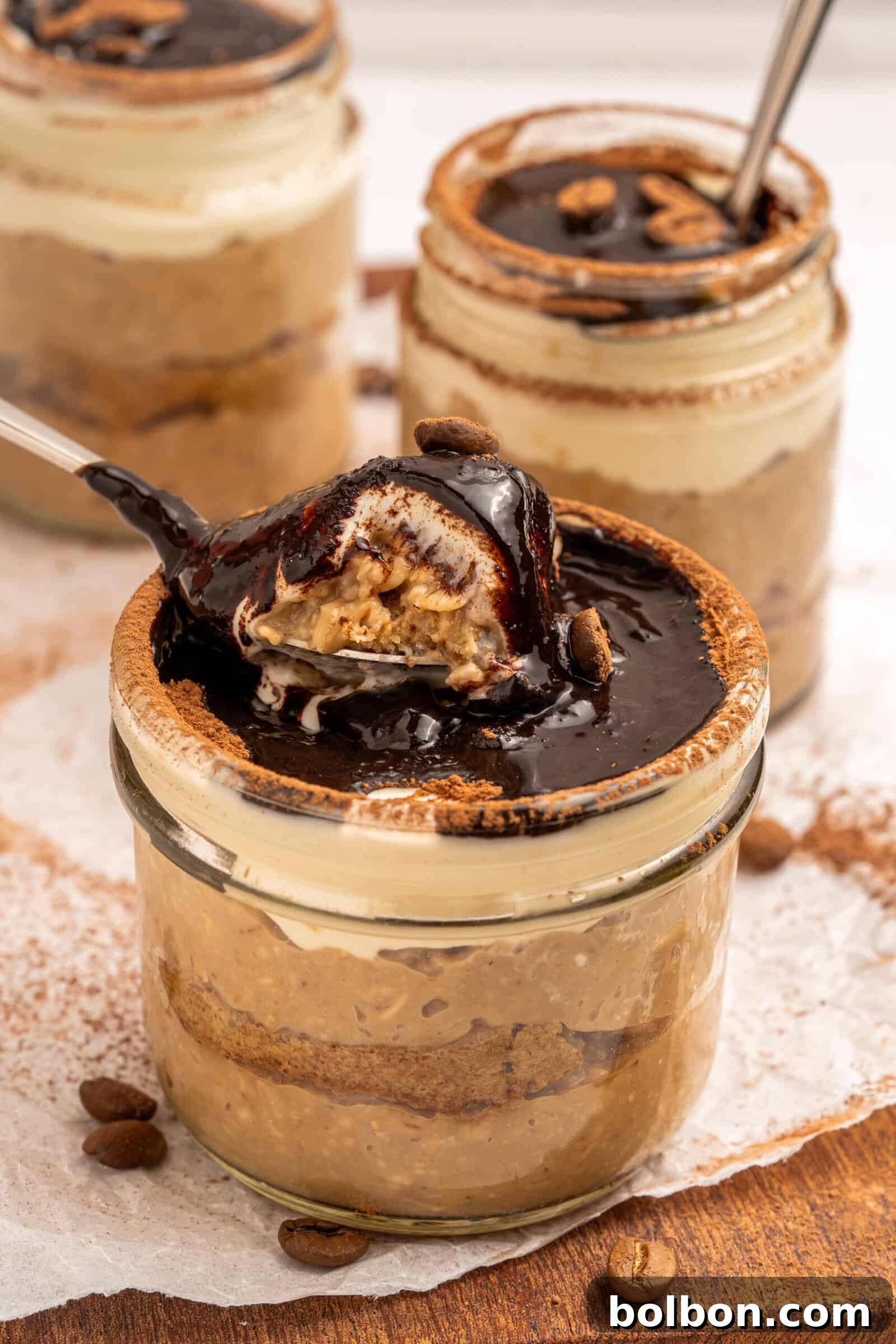 Three glass jars filled with creamy tiramisu, topped with chocolate sauce and coffee beans. One jar has a spoonful lifted, showing layers of dessert inside. The jars rest on parchment paper with scattered coffee beans.