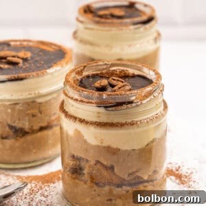 Three jars of tiramisu dessert layered with cream, cake, and cocoa, topped with chocolate sauce and coffee beans, sit on a wooden board with cocoa powder and coffee beans scattered around.