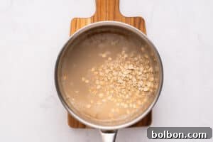 A metal saucepan with oats soaking in water sits on a small wooden cutting board, viewed from above on a white surface.