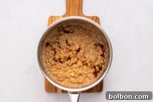 A saucepan filled with cooked oatmeal, topped with a swirl of dark syrup, sits on a small wooden cutting board against a light-colored background.