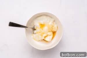 A white bowl with plain yogurt and honey, partially mixed, with a metal spoon resting inside, on a white surface.