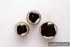Three small glass jars filled with a creamy dessert topped with a layer of glossy dark chocolate sauce, arranged in a triangle on a white marble surface.