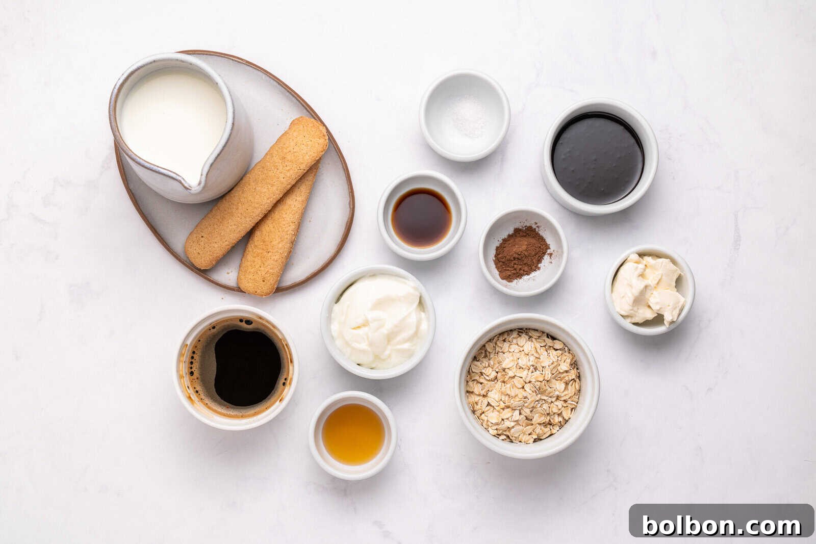 Assorted ingredients in small bowls on a white surface, including milk, ladyfinger biscuits, coffee, oats, yogurt, cocoa powder, vanilla, syrup, mascarpone cheese, salt, and sweetener.