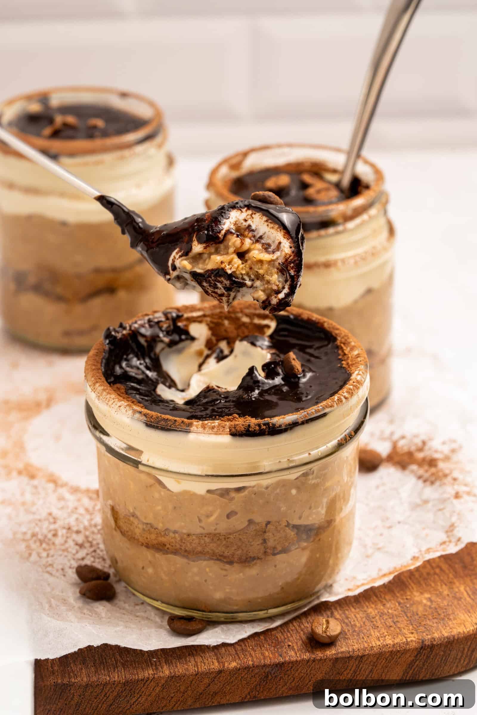 Three glass jars filled with layered tiramisu are topped with chocolate sauce and whipped cream. A spoon is lifting a portion from the front jar, revealing the creamy, layered inside. The jars sit on a wooden board with coffee beans.