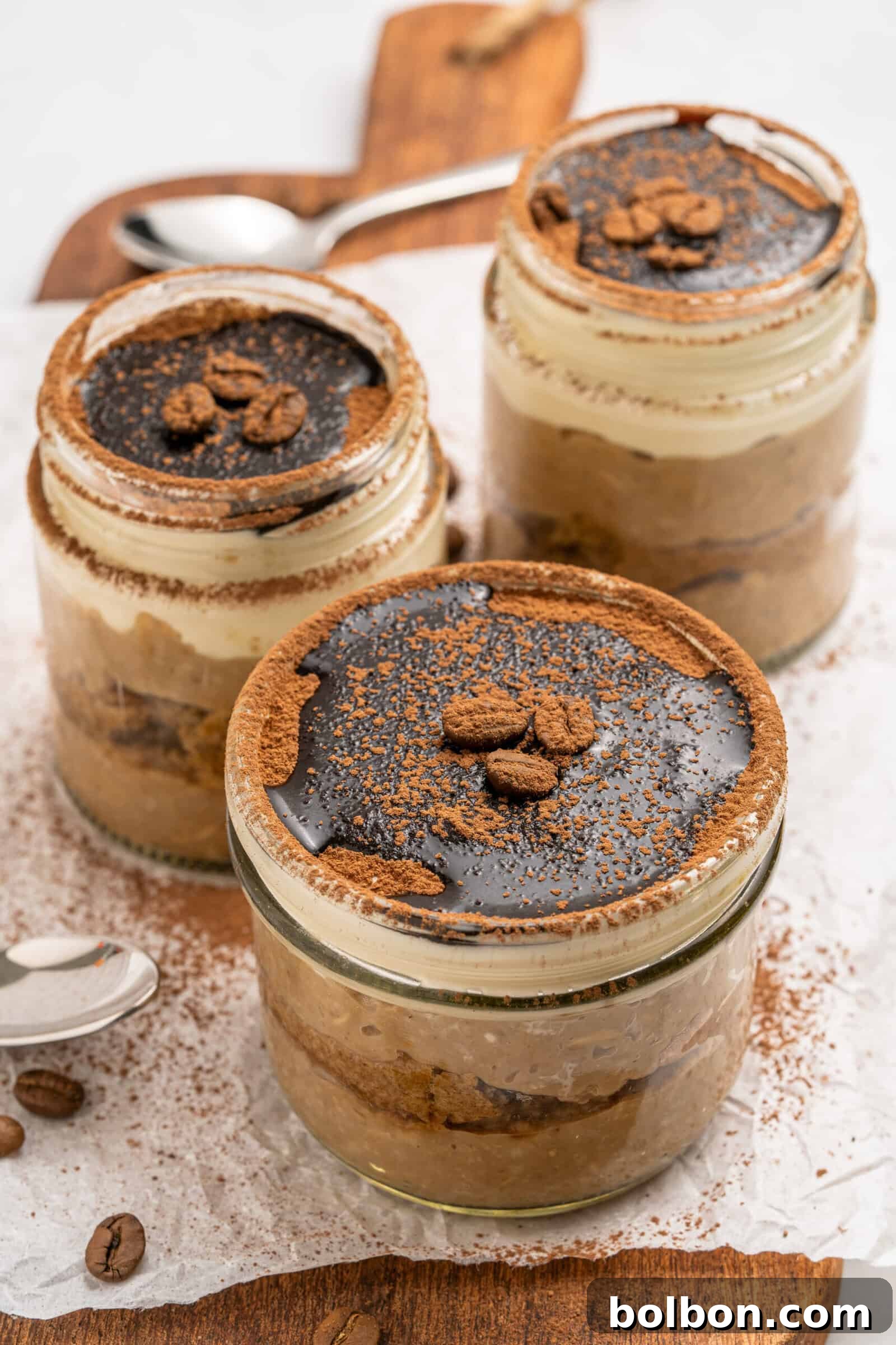 Three glass jars filled with layered tiramisu dessert, topped with cocoa powder, chocolate, and coffee beans, are arranged on a wooden board with a spoon and scattered coffee beans nearby.