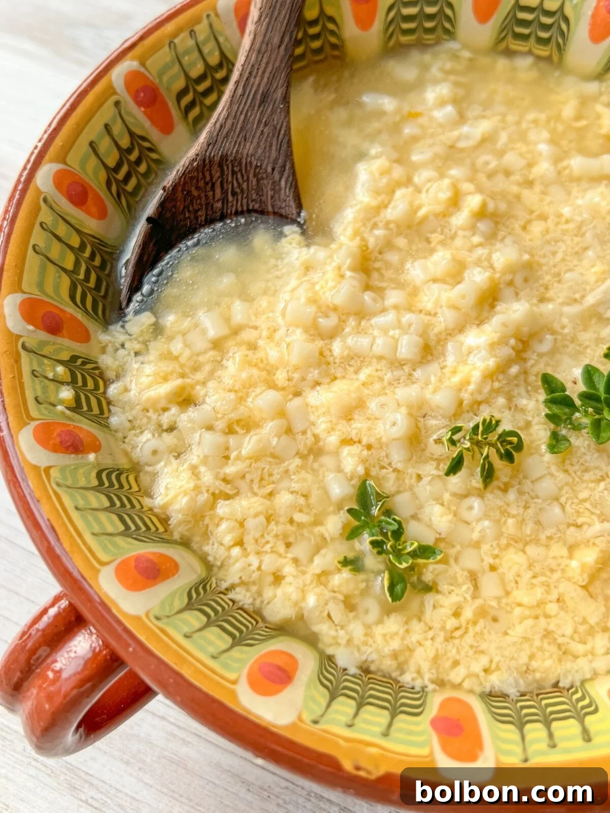 A close-up of a bowl of soup with small pasta and cheese. The soup is garnished with a few sprigs of fresh herbs. The bowl has a vibrant, colorful pattern with red and yellow tones. A wooden spoon rests in the soup.
