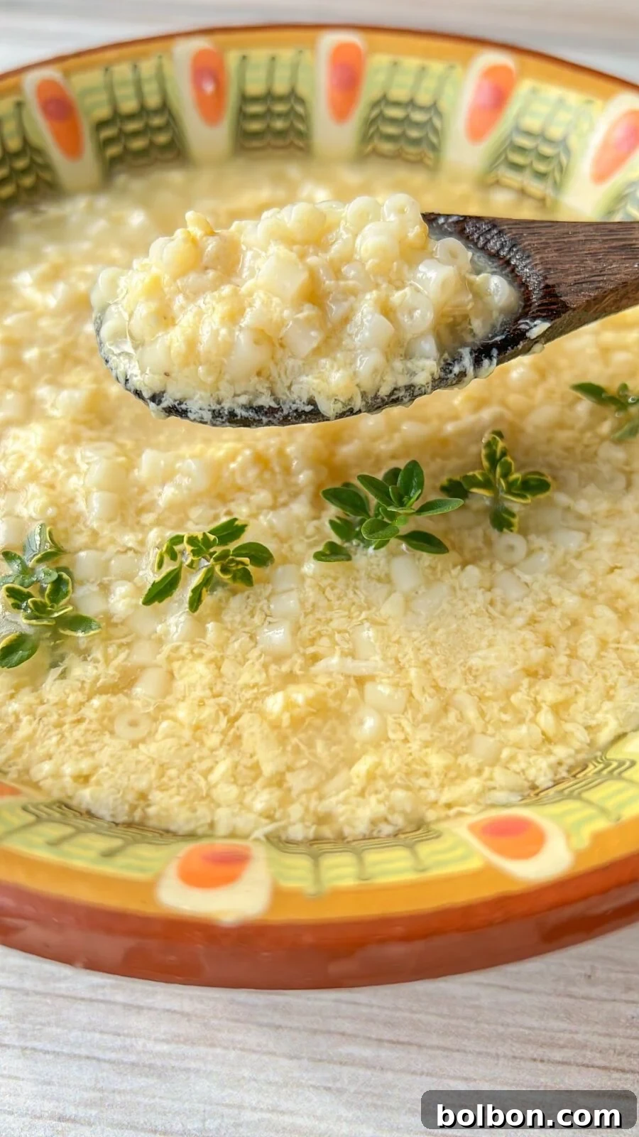 A close-up of a bowl of Stracciatella Soup garnished with green herbs. The soup contains small, round grains and is served in a decorative ceramic bowl. A wooden spoon is lifting a portion of the grains from the soup.