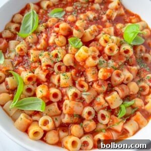 A white bowl filled with ditalini pasta in a rich tomato sauce, Pasta e Ceci, garnished with fresh basil leaves.