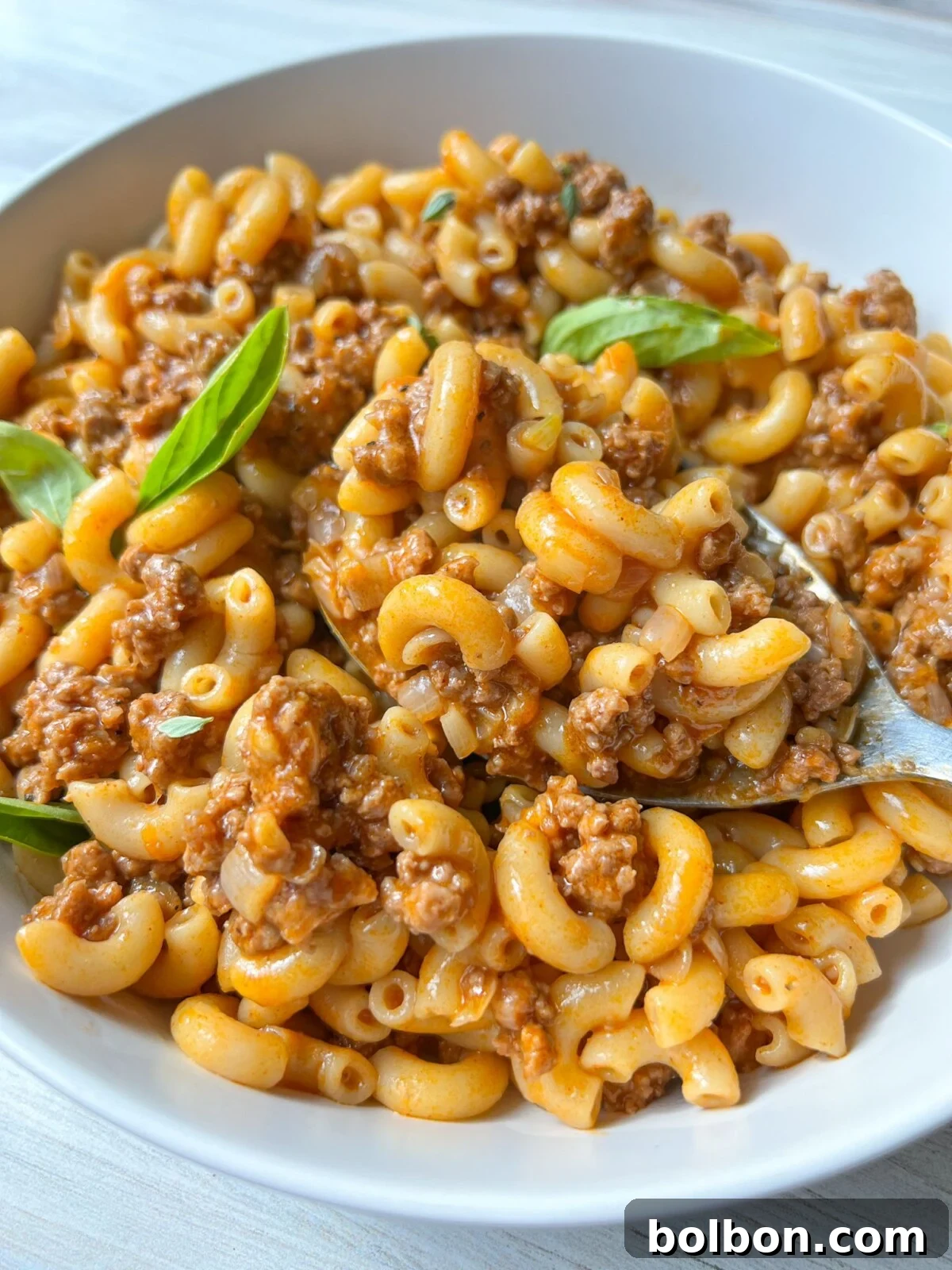 A white bowl filled with cooked elbow macaroni mixed with a meaty tomato sauce. A few fresh basil leaves garnish the dish. A spoon is partially visible, resting in the macaroni, which looks flavorful and hearty.