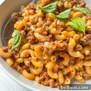 A close-up of macaroni pasta mixed with ground meat in a tomato-based sauce, reminiscent of classic Hamburger Helper, garnished with fresh basil leaves and served in a white bowl with a spoon.