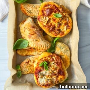 A tray lined with parchment paper holds mini pizzas topped with cheese and herbs, and several calzones. Fresh basil leaves are scattered on top for garnish.
