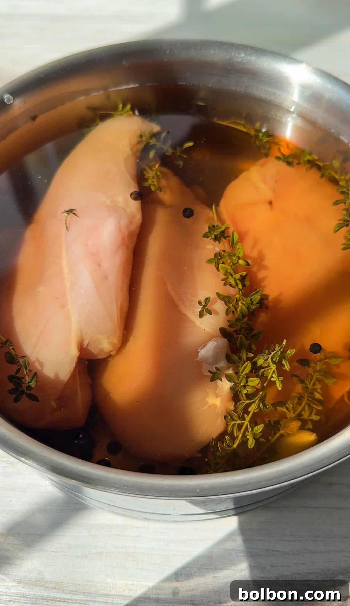 Raw chicken breasts soaking in a stainless steel bowl filled with water, peppercorns, and fresh thyme sprigs. The bowl is placed on a light-colored surface with sunlight streaming over it.