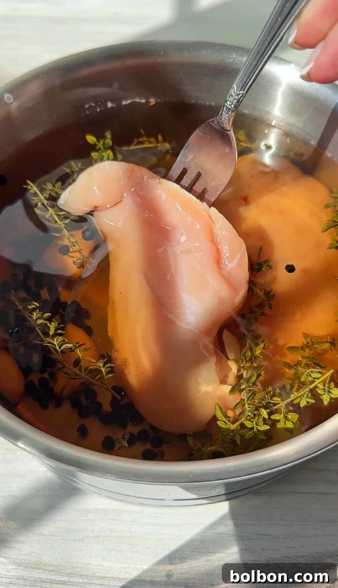 A person uses a fork to lift a raw piece of chicken breast from a bowl filled with water, herbs, and peppercorns. The scene is brightly lit, highlighting the ingredients in the bowl for this Chicken Deli Meat Recipe.