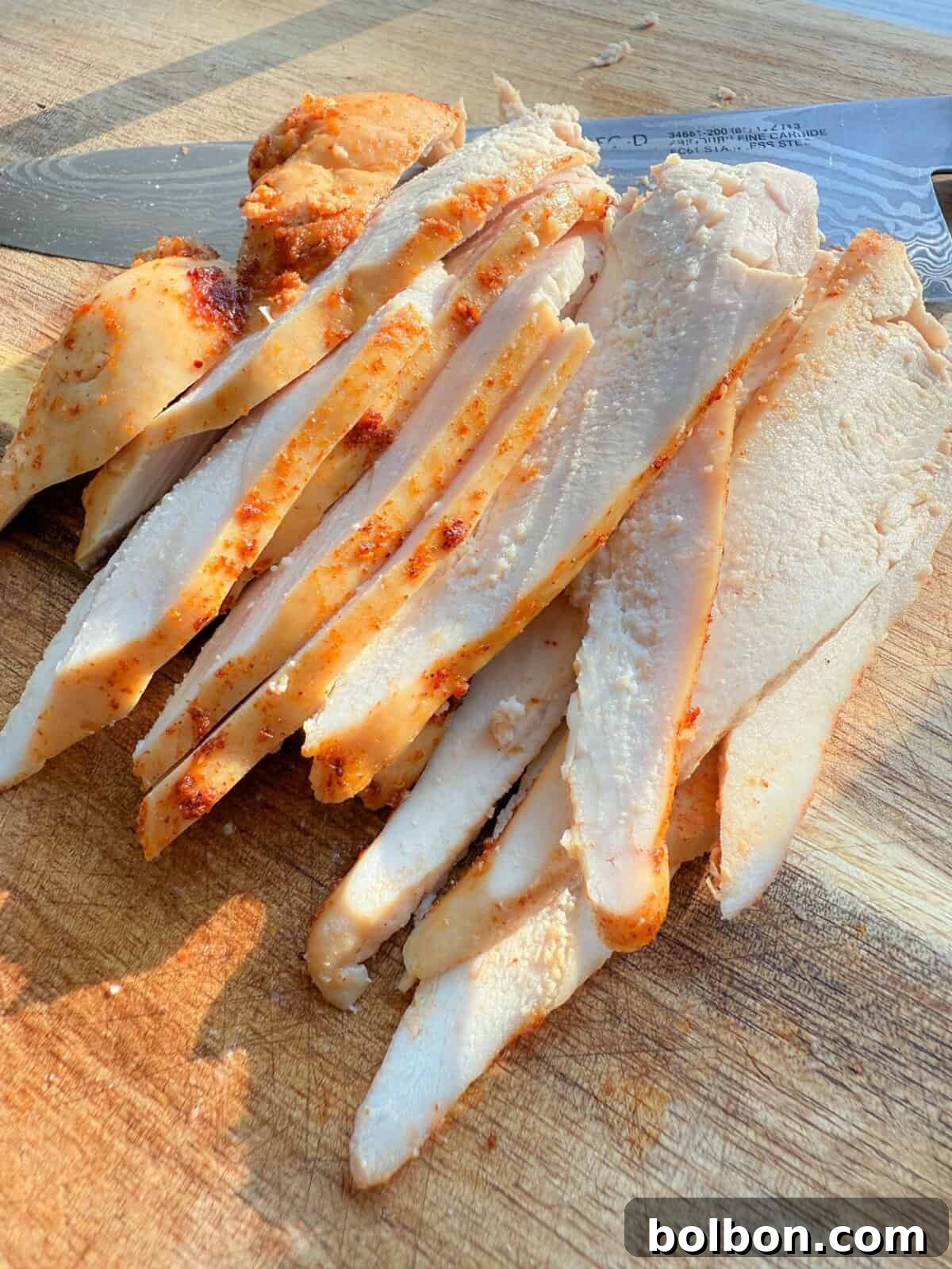 Sliced cooked chicken breast on a wooden cutting board, with a knife in the background. The chicken is seasoned with spices, giving it a slightly red and browned exterior. The slices for this Chicken Deli Meat Recipe are thin and arranged neatly.