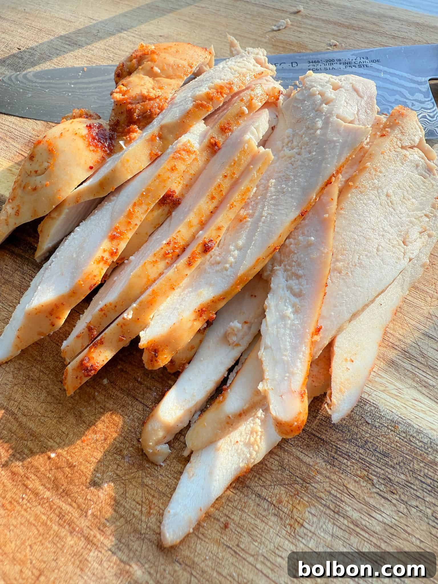 Sliced Chicken Deli Meat arranged on a wooden cutting board with a knife in the background. The chicken appears to be seasoned and cooked, showing a slightly golden-brown exterior and juicy interior. The board has a textured surface with visible grain patterns.