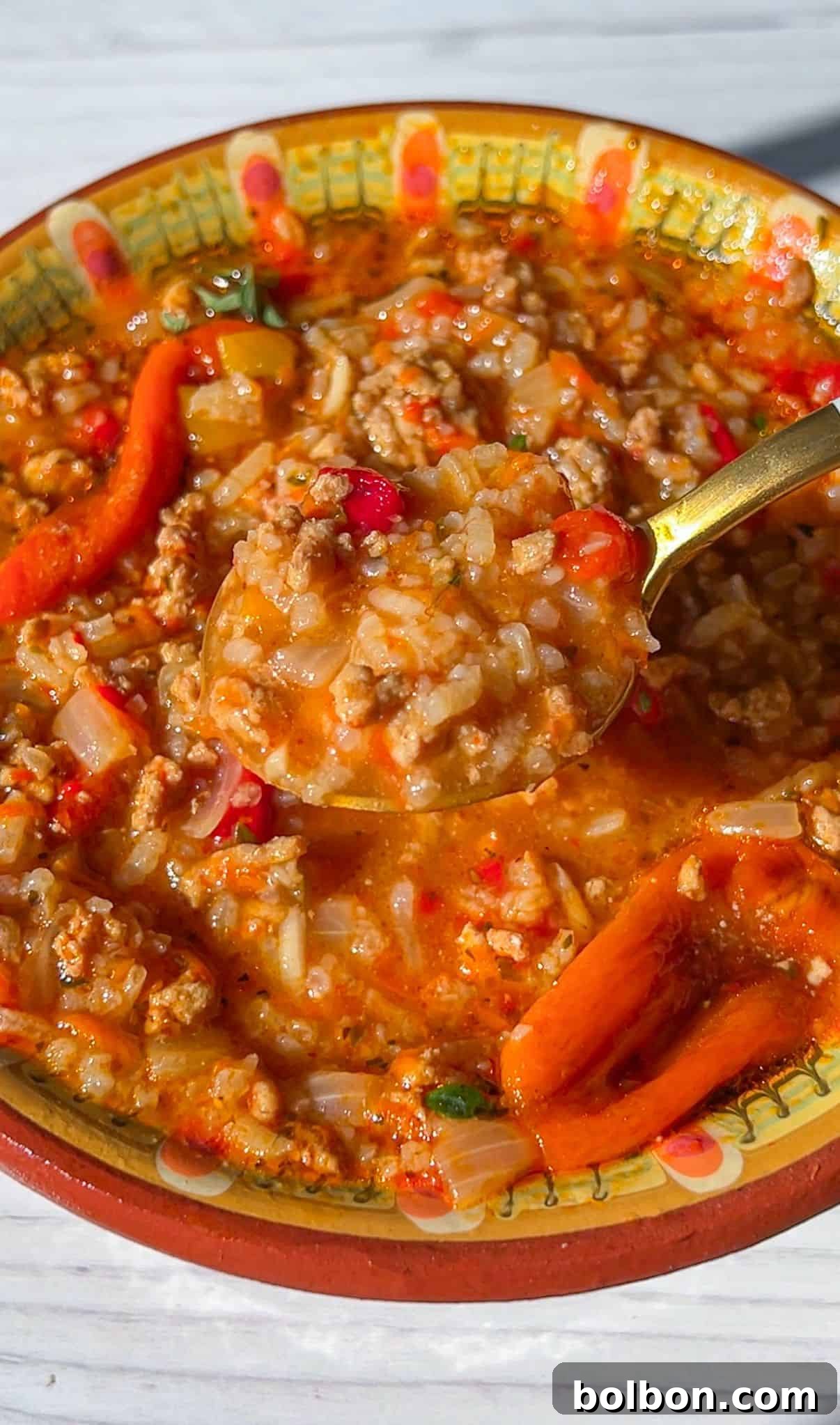 A colorful and hearty bowl of Stuffed Pepper Soup, featuring ground meat, tender rice, and vibrant red pepper pieces. A spoon delves into the chunky texture, highlighting the rich colors. The inviting dish is presented in a beautifully decorated ceramic bowl, promising warmth and comfort.