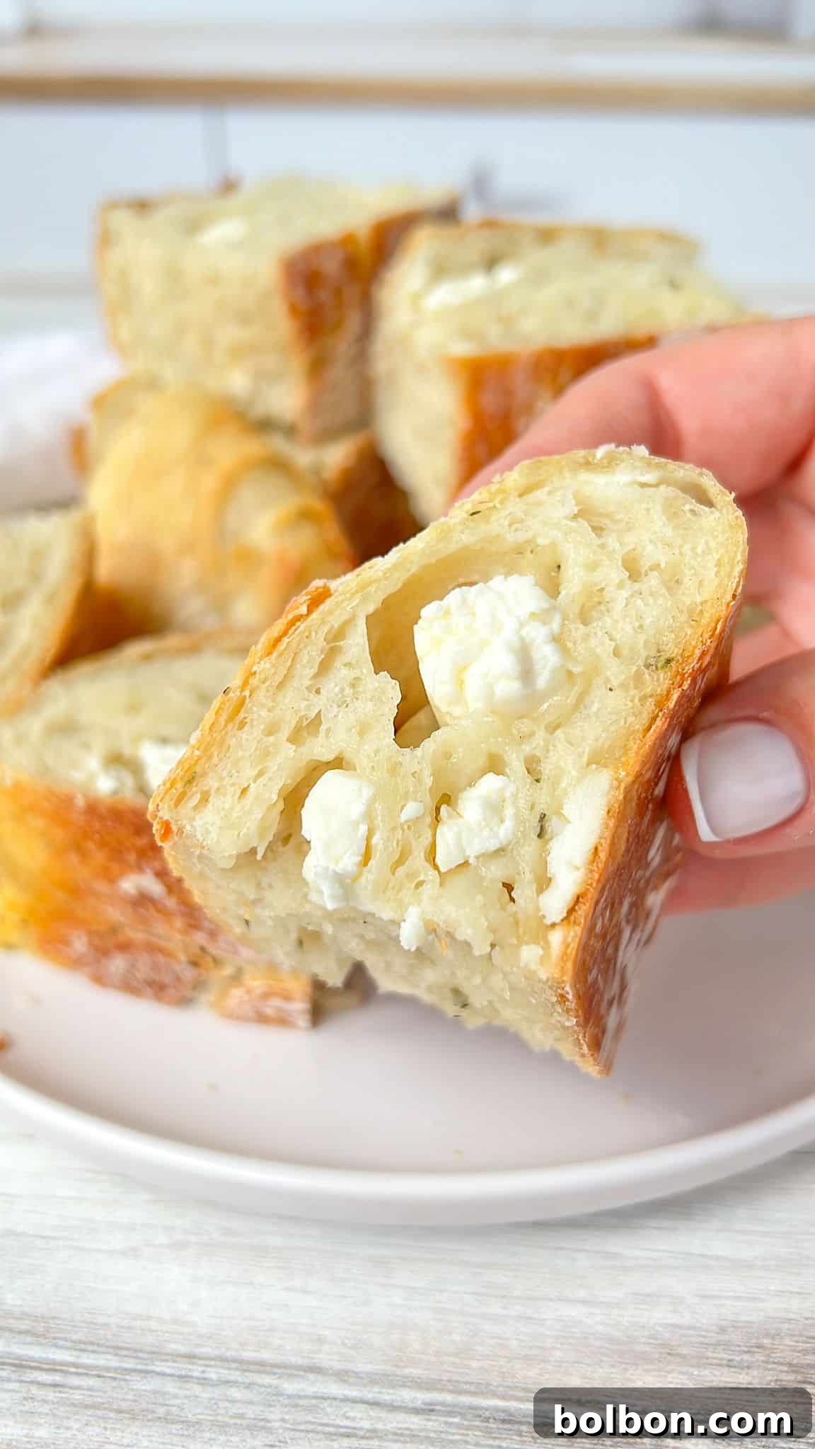 A hand holding a piece of rustic no knead feta bread with visible chunks of cheese, set on a white plate with more bread pieces in the background.