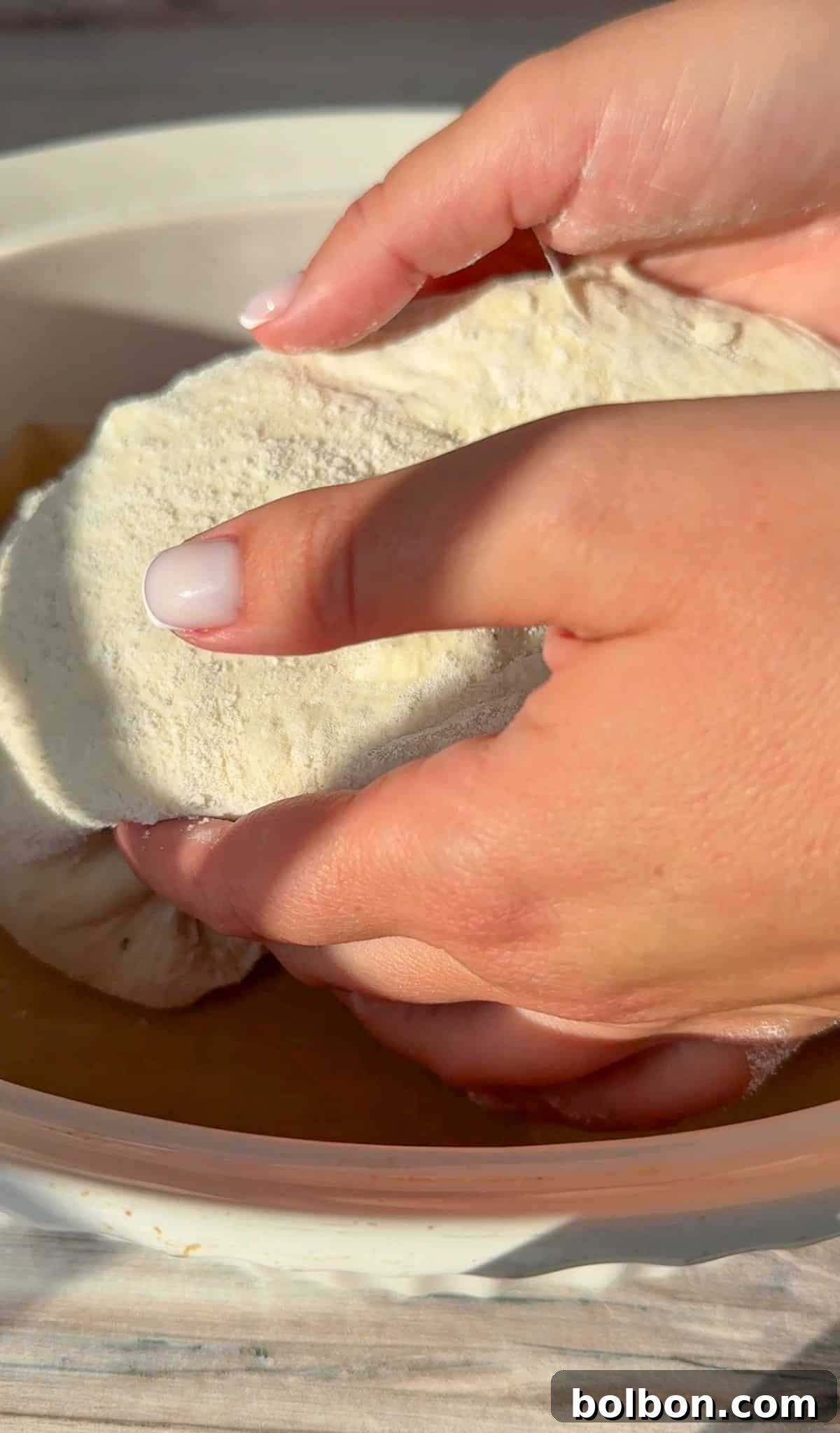 Close-up of hands placing the no knead feta bread in to a sunlit bowl. The focus is on the white flour-dusted dough being worked with care. The hands have manicured nails, and the scene suggests baking in progress.