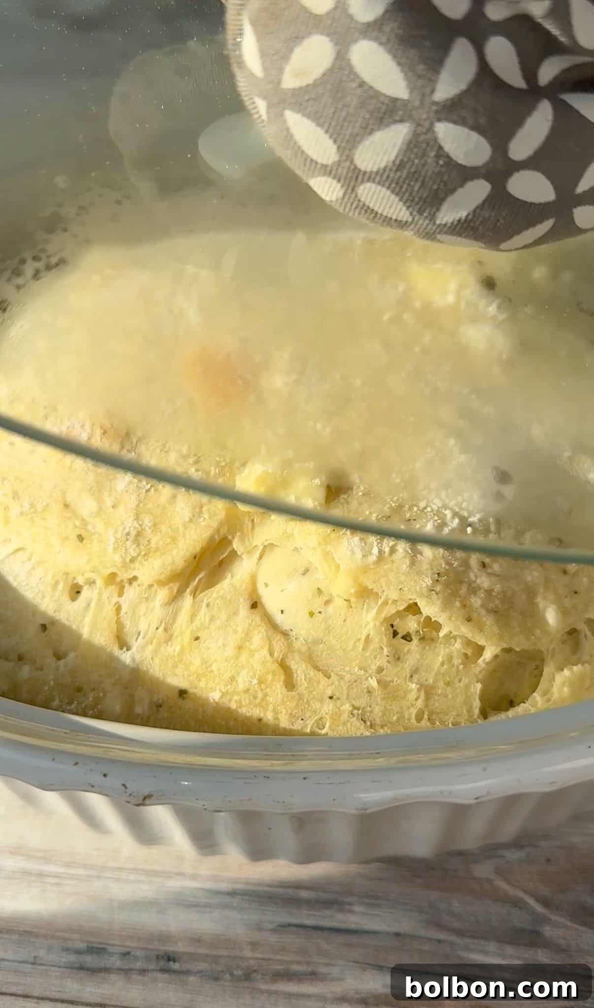 A close-up of a steamy no knead feta bread being cooked under a glass lid. The dish appears to be a fluffy, baked meal with visible chunks, possibly cheese or vegetables. A hand holding a patterned oven mitt touches the lid.