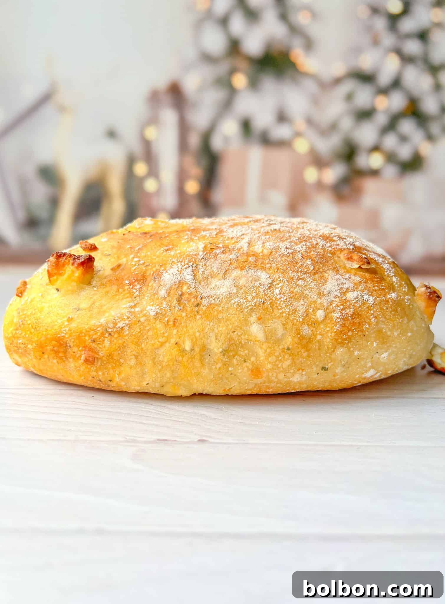 A freshly baked, golden-brown loaf of no knead feta bread bread dusted with flour sits on a light wooden surface. The background is softly blurred with hints of festive decorations, including lights and greenery.