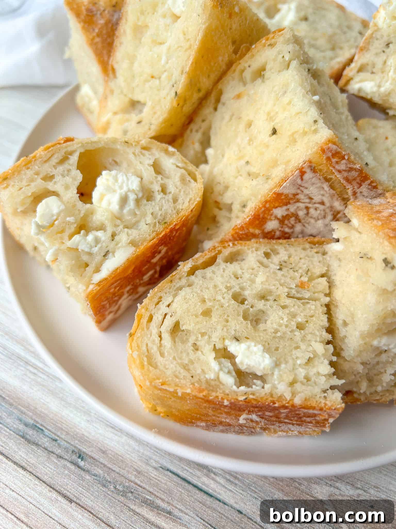 A plate filled with slices of crusty no knead feta bread, featuring a golden-brown crust and soft, airy interior. The bread has visible pockets of cheese, adding texture and flavor. The plate is set on a light wooden surface.