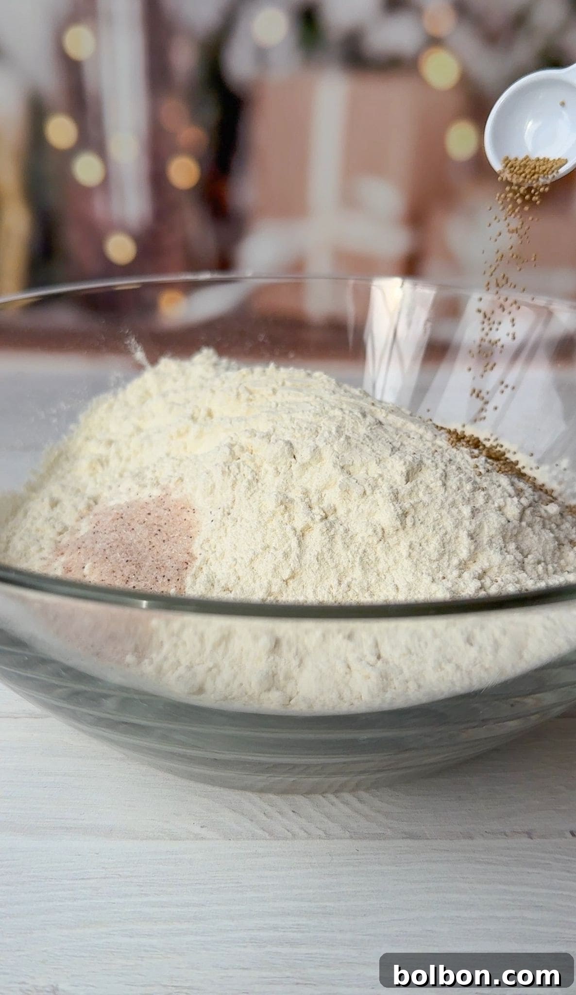 A clear glass bowl filled with flour and other dry ingredients. A spoon is pouring yeast into the bowl. There are blurred holiday decorations and wrapped presents in the background.