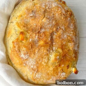 A freshly baked round loaf of bread with a golden, crispy crust rests on a light cloth on a white wooden surface. The bread is dusted with flour, showcasing its rustic texture.